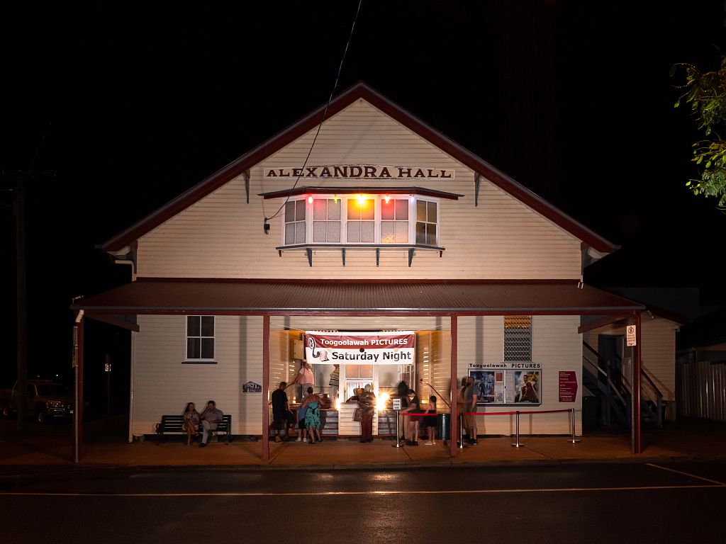 Outside the Toogoolawah Pictures at Alexandra Hall, An old style Hall with a box office out the front. Photo is taken at night with people waiting to buy tickets