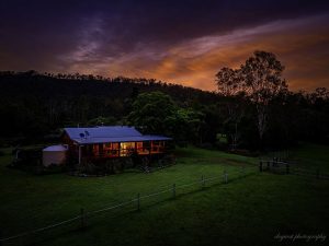 The Log Cabin at Mount Byron Retreat. Photo has been taken in the early evening as its getting dark with lights on. The cabin is surrounded by green grass and trees with a mountain silhouette in the background
