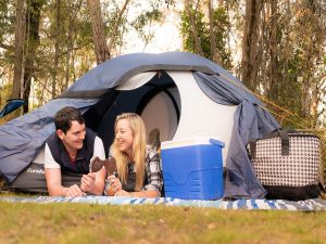 Couple leaning out of the pop up tent eating ice creams at Jimna Base Camp. Bushland setting behind them. Esky and picnic blanket in front of the blue and white tent.